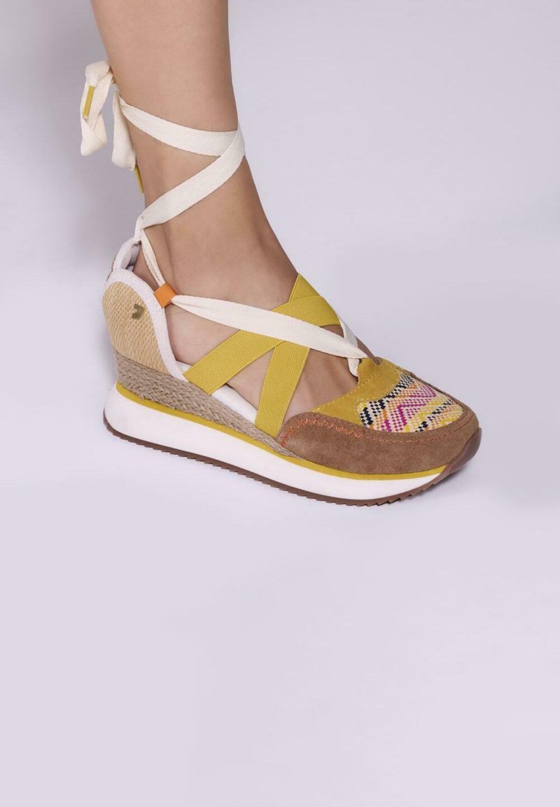Baskets compensées avec un bout en daim marron, des accents jaunes, un panneau latéral tissé et des brides blanches croisées. Présente un détail coloré et patterned sur le devant.