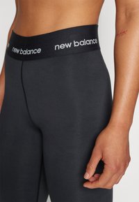 Juodos leggingai su plačiu juosmeniu, kuriame matomas "new balance" logotipas. Medžiaga atrodo lygi, o dizainas yra prigludęs ir su minimaliais siūlais.