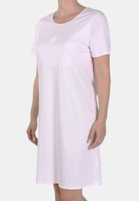 Donna che indossa un vestito semplice rosa chiaro, con maniche corte e lunghezza fino al ginocchio, dotato di un colletto rotondo e un sottile emblema circolare sul petto.