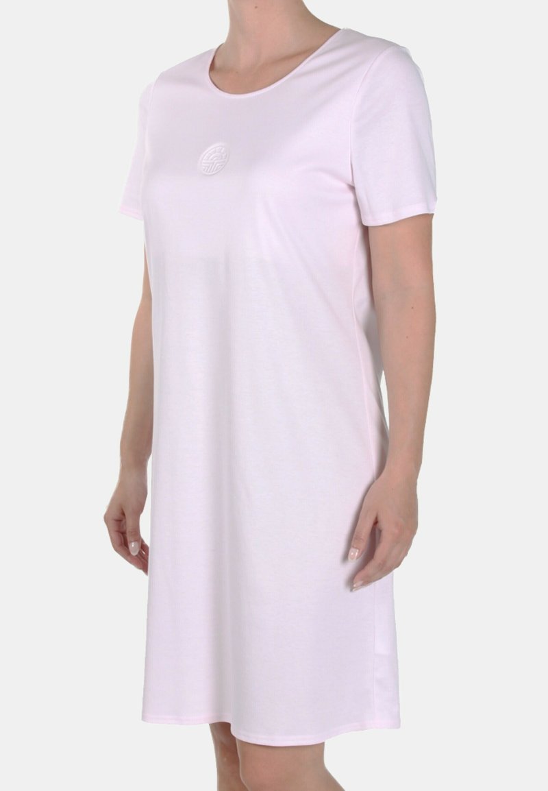 Donna che indossa un vestito semplice rosa chiaro, con maniche corte e lunghezza fino al ginocchio, dotato di un colletto rotondo e un sottile emblema circolare sul petto.