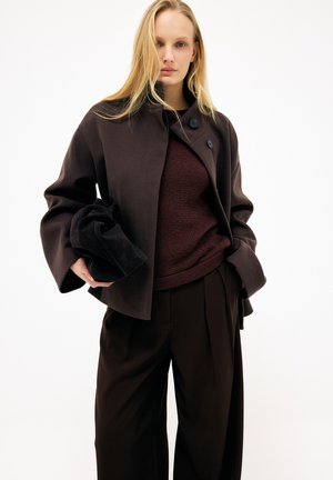 Donna che indossa un cappotto oversize marrone scuro, maglione lavorato a maglia color granata, pantaloni neri a gamba larga, che tiene una pochette nera contro uno sfondo bianco.