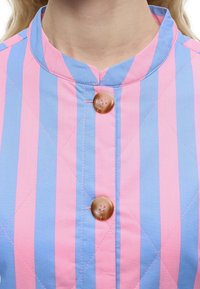 Κοντινό πλάνο ενός ατόμου που φοράει πουκάμισο με κατακόρυφες ρίγες σε ροζ και μπλε χρώμα, με όρθιο γιακά και δύο καφέ κουμπιά κουμπωμένα.