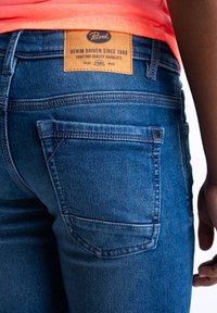 Jeans de mezclilla azul con un bolsillo trasero, que cuentan con una etiqueta de marca de cuero en la cintura. La tela tiene un ligero desvanecimiento y detalles de costura.