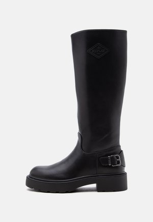 Botte haute en cuir noir avec semelle épaisse, détail de boucle latérale, et patch logo "Replay" près du haut sur fond blanc uni.