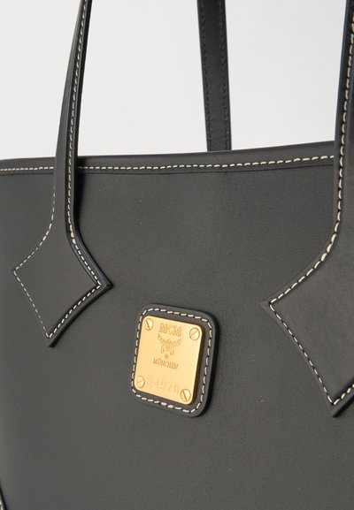 Μαύρη δερμάτινη τσάντα χειρός με αντίθετη λευκή ραφή και χρυσή πλακέτα με το λογότυπο MCM München στο μπροστινό μέρος κάτω από τις δύο λαβές.
