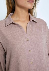 Light pink button-up shirt in a linen blend with a collar, featuring a textured surface and five small brown buttons down the front.