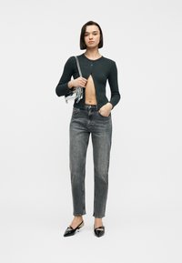 Γυναίκα με σκούρο κοντό ζακέτα, ξεθωριασμένο μαύρο τζιν, μαύρες μυτερές γόβες και κρατώντας διάφανη τσάντα ώμου μπροστά σε απλό φόντο.