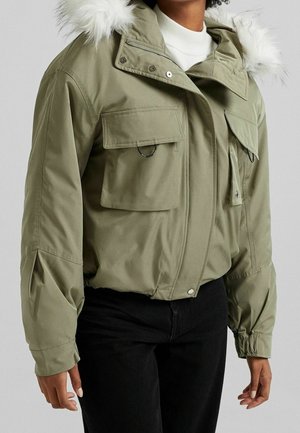 Krótkja kurtka oliwkowa z dużymi kieszeniami z patkami, czarnymi pierścieniami, elastycznymi mankietami oraz białym sztucznym futerkiem przy kapturze, noszona na białej bluzce.