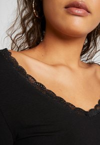 Ausschnitt einer Frau mit schwarzem Oberteil mit Spitzenbesatz, lockigem Haar, goldenen Creolen und sichtbar natürlichem Lippenstift.