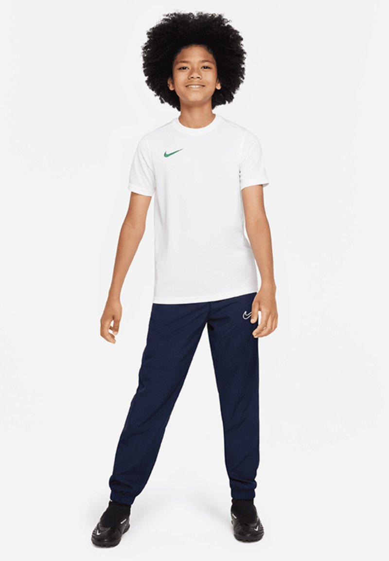 T-shirt Nike bianco con swoosh verde, abbinato a pantaloni sportivi blu navy. Tessuto texture, maniche corte, vita elastica e scarpe nere.