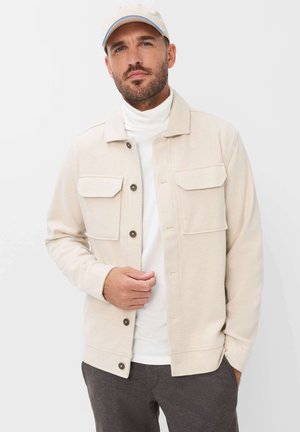 Mand iført en beige jakke med knapper og klaplommer, en hvid rullekrave, en beige kasket og mørke bukser, stående mod en ensfarvet hvid baggrund.