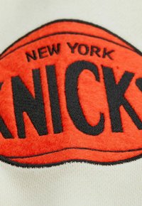 Rot und schwarz gesticktes "NYC KNICKS"-Logo auf einem hellen, strukturierten Stoff mit weichen, filzartigen Details im kreisförmigen Design.