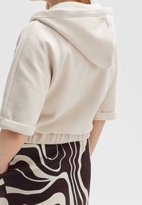 Světle béžová cropped mikina s ohrnutým manžetem a kapucí na šňůrku, vyrobená z měkkého materiálu, v kombinaci s kostkovanou sukní v černobílé barvě.