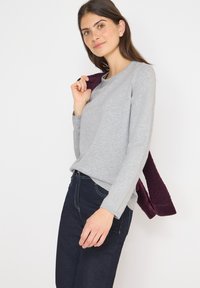Maglietta grigia a maniche lunghe con scollatura rotonda, indossata con jeans scuri. Un maglione bordeaux a trama è appoggiato su una spalla.