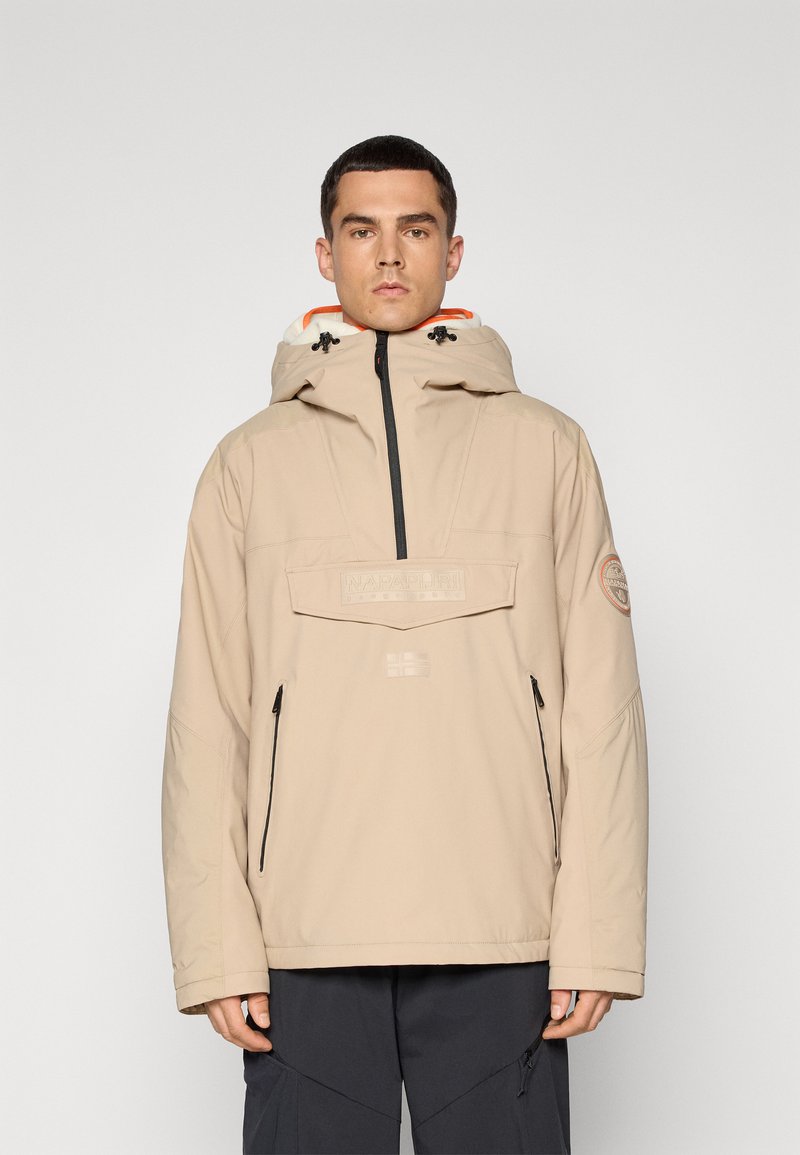 Veste à capuche beige avec des zips noirs, présentant une coupe oversized, une poche avant et une doublure orange. Écusson logo sur la manche gauche.