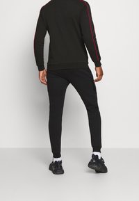 Uomo in piedi di spalle, che indossa una tuta nera con strisce rosse sulle maniche, scarpe da ginnastica nere e calzini bianchi, su uno sfondo neutro.