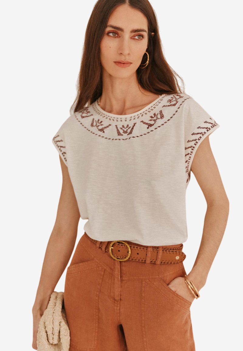 Haut gris clair à manches courtes avec des détails brodés autour de l'encolure, associé à un pantalon taille haute de couleur rouille et une ceinture tressée.