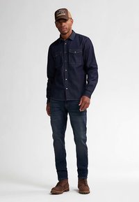 Camicia in denim scuro con due tasche sul petto, abbinata a jeans scuri e stivali in pelle marrone. La persona indossa un berretto da baseball color beige.