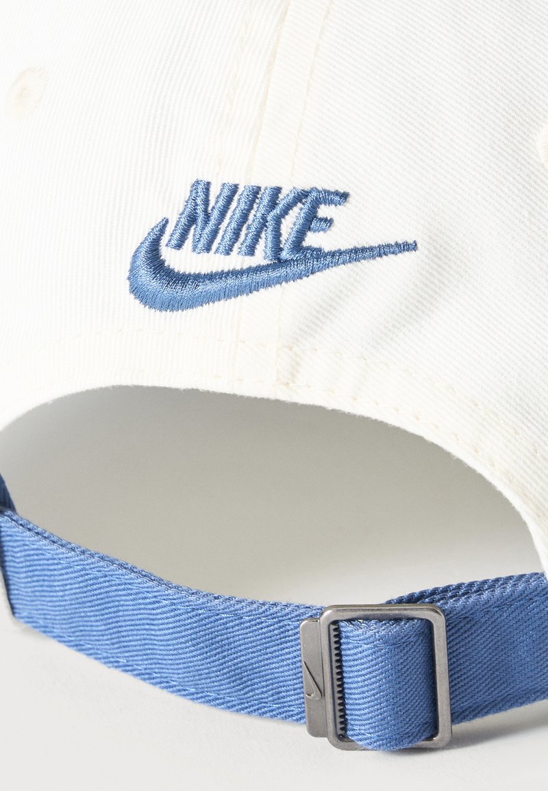 Weiße Stoffkappe mit blau gesticktem Nike-Logo und verstellbarem blauem Riemen mit Metallschnalle.