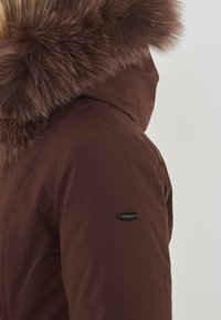 Giacca isolante marrone con un ampio cappuccio in pelliccia soffice e un logo in tessuto sulla manica sinistra. Tessuto liscio con un design aderente.