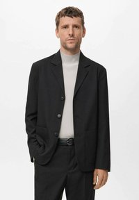 Blazer nero con rever a punta, due tasche frontali e tre bottoni. Indossato sopra un dolcevita a coste grigio chiaro. Vestibilità tailored.