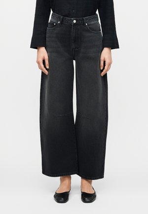 Άτομο που φοράει μαύρο τζιν παντελόνι με φαρδιά μπατζάκια, σε συνδυασμό με μαύρα επίπεδα παπούτσια και μακρυμάνικο μαύρο τοπ, με φόντο έναν απλό τοίχο.