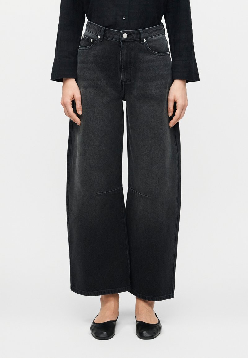 Άτομο που φοράει μαύρο τζιν παντελόνι με φαρδιά μπατζάκια, σε συνδυασμό με μαύρα επίπεδα παπούτσια και μακρυμάνικο μαύρο τοπ, με φόντο έναν απλό τοίχο.