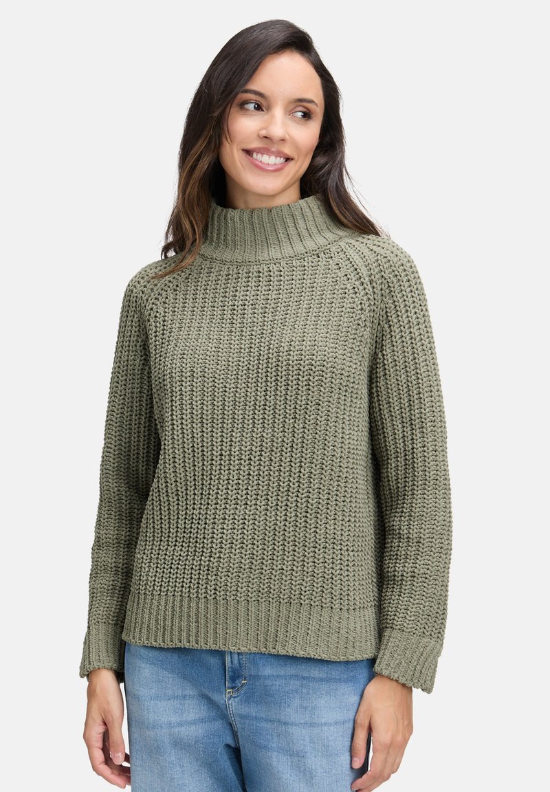 Kobieta uśmiechająca się z boku, ubrana w grubą olive green sweter z golfem i jasnoniebieskie dżinsy, na jasnym tle.