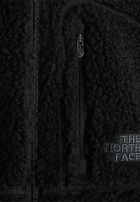 Μαύρο fleece ύφασμα με δύο κάθετα μαύρα φερμουάρ, ένα μικρό με μεταλλικό τράβηγμα με λογότυπο και το λογότυπο "The North Face" κεντημένο με μπλε κλωστή.