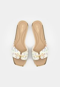 Sandales blanches à enfiler avec motif floral, garnitures dorées, forme à bout carré et semelle intérieure beige rembourrée. Logo de la marque visible sur la semelle intérieure.