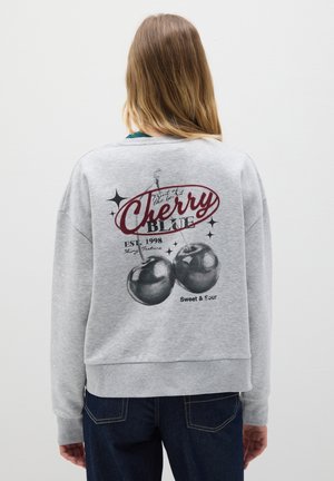 Donna con capelli lunghi che indossa una felpa grigia con una grafica di ciliegie, testo "Cherry Blue," e jeans scuri, rivolta di spalle su uno sfondo bianco.