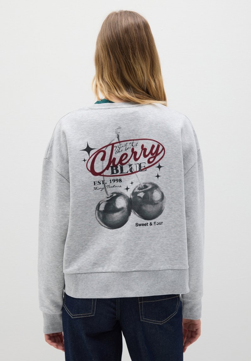 Donna con capelli lunghi che indossa una felpa grigia con una grafica di ciliegie, testo "Cherry Blue," e jeans scuri, rivolta di spalle su uno sfondo bianco.