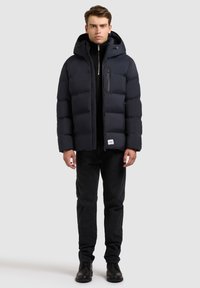 Tengerészkék pufi dzseki kapucnival, varrott panelekével, cipzáras elülső zsebével és állítható húzózsinórral. Fekete farmernadrággal és csizmával párosítva.