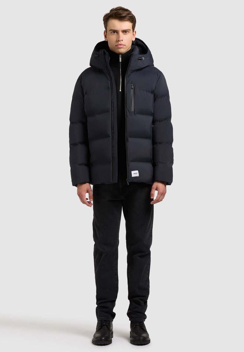 Tengerészkék pufi dzseki kapucnival, varrott panelekével, cipzáras elülső zsebével és állítható húzózsinórral. Fekete farmernadrággal és csizmával párosítva.