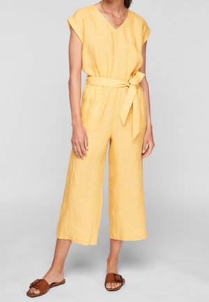 Kvinde iført en gul ærmeløs jumpsuit med taljebånd og brede cropped ben, kombineret med brune flade slip-in sandaler.