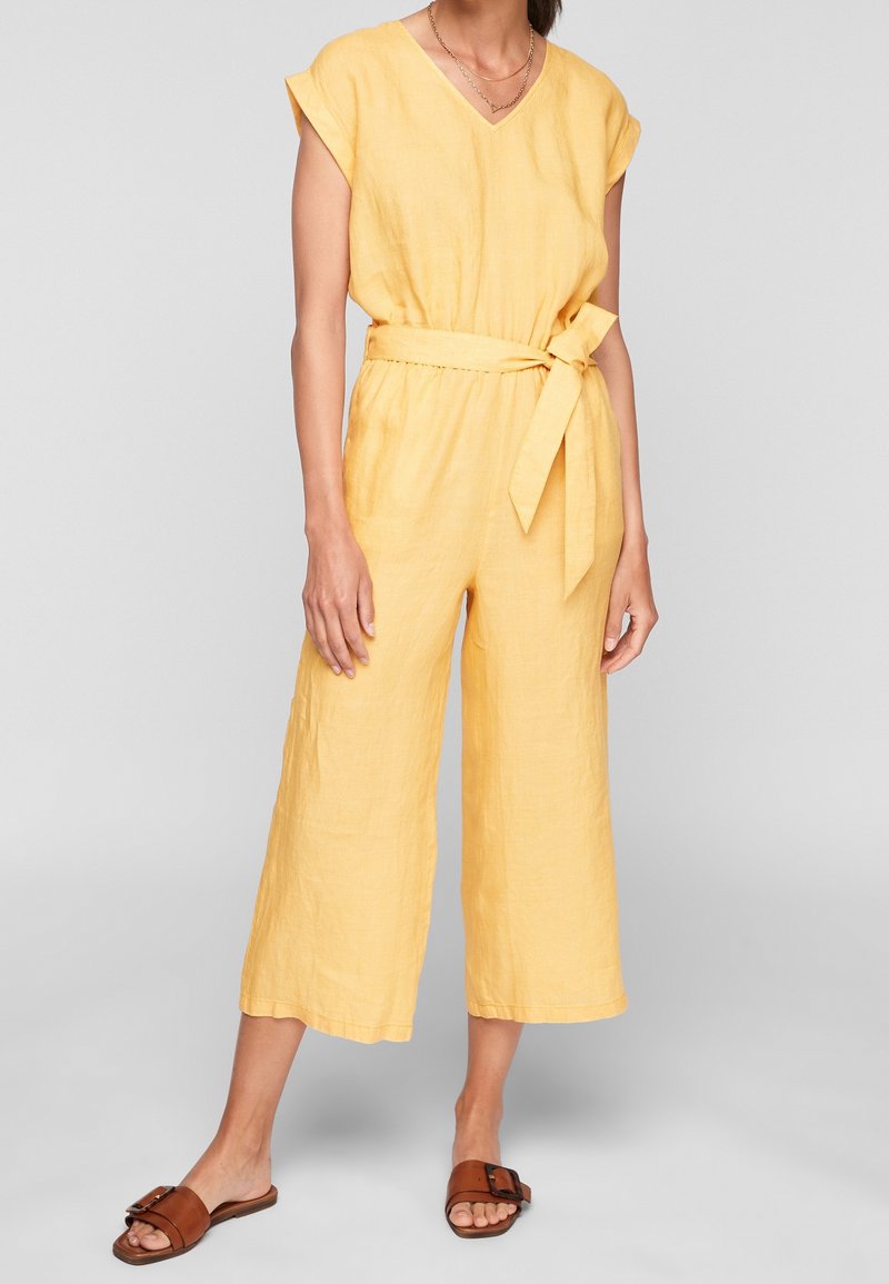 Femme portant une combinaison jaune sans manches avec une ceinture à la taille et des jambes larges et coupées, associée à des sandales plates marron à enfiler.