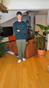 Πράσινη φόρμα, μπλε σκούρο φούτερ με λογότυπο, λευκά αθλητικά παπούτσια. Στέκεται σε σαλόνι με καφέ ριμπ καναπέ και μεγάλο πράσινο φυτό.