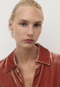 Modello che indossa una camicia button-up in velluto color ruggine con una texture morbida e colletto a punta, dotata di cinque bottoni sul davanti.