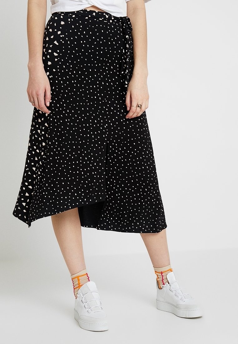 Jupe midi portefeuille noire avec petits points blancs irréguliers, portée avec des baskets blanches et des chaussettes rayées colorées sur une personne debout.