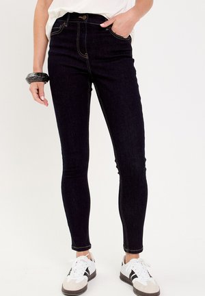 Jean skinny en denim noir à taille haute, avec des coutures dorées, des poches avant et une fermeture à bouton classique. Associés à des baskets blanches.