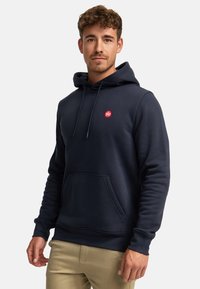Sudadera de color azul marino hecha de una tela suave, con un bolsillo canguro en la parte frontal y un pequeño logo rojo en el lado izquierdo del pecho. Capucha ajustable con cordón.