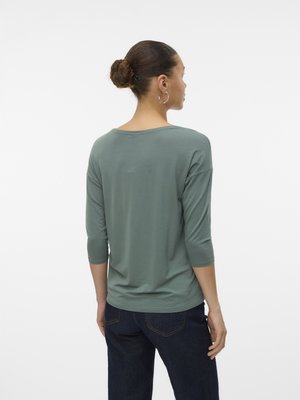 Bun hajú nő ezüst karika fülbevalót visel, zöld háromnegyedes ujjú felsőben és sötétkék farmernadrágban áll háttal egy fehér háttér előtt.