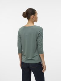 Femeie cu coc, purtând cercei rotunzi din argint, un top verde cu mânecă trei sferturi și blugi de culoare bleumarin, întoarsă cu fața spre spate pe un fundal alb.