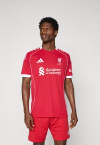 Tricou de fotbal roșu cu accente albe, având logo-ul Liverpool FC și logo-ul sponsorului "Standard Chartered". Mâneci scurte, material ușor.