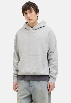 Un hoodie gris en matériau doux avec une grande poche avant, un capuchon à cordon et des poignets. Porté par-dessus une chemise à couches gris foncé.