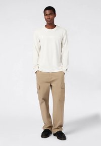 Camicia crema a maniche lunghe con logo "Champion", abbinata a pantaloni cargo beige con molteplici tasche laterali e scarpe nere.