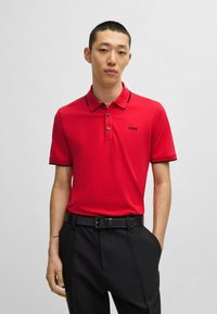 Rød polo skjorte med sorte accenter og logo, kortærmet og med krave. Bæres med sorte bukser og et bælte. Glat stof, figursyet design.