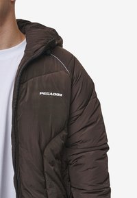 Brun puffjacka med huva, med dragkedja, stickade mönster och "PEGADOR"-logotyp i vitt på bröstet.