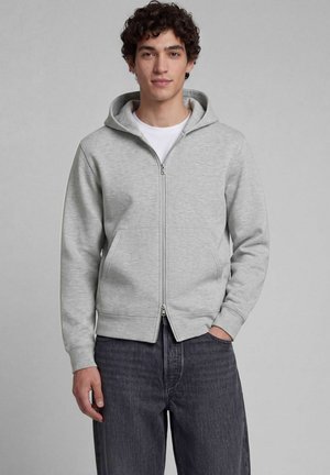 Giovane uomo con capelli ricci e scuri che indossa una felpa con cappuccio grigio chiaro con zip sopra una camicia bianca e jeans grigio scuro su uno sfondo semplice.