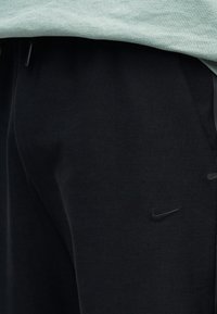 Calças atléticas pretas com textura suave, apresentando um logotipo sutil da Nike em preto na lateral e detalhes cinzentos nos bolsos.
